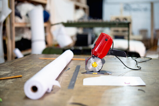 A red electric fabric cutting machine placed on a wooden work table with fabric roll and sewing patterns in a garment workshop.