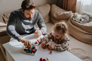 A 3-year-old girl and her father play with modeling clay in a sunny living room. Unfiltered,...
