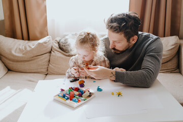 Naklejka premium A 3-year-old girl and her father play with modeling clay in a sunny living room. Unfiltered, authentic photography capturing real family moments of creativity and bonding