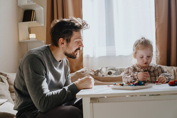 A 3-year-old girl and her father play with modeling clay in a sunny living room. Unfiltered,...