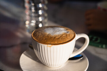 A white ceramic cup of frothy cappuccino sits on a saucer in soft natural light, evoking cozy café vibes.