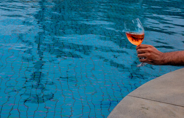 Hand holding glass of rosé wine near swimming pool edge, relaxed summer leisure moment,