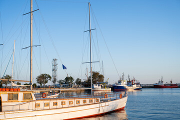 Fototapeta premium A wooden sailboat with white hull rests in a calm harbor, surrounded by colorful ships and reflected in tranquil water.