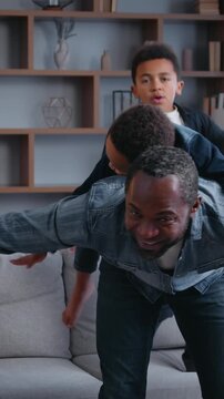Happy African American family playing piggyback at home joyful carefree father dad riding on back little son spinning play with two kids boys sons children having fun fooling around jumping on couch