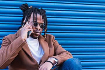 Stylish African man talking on phone while leaning on blue wall