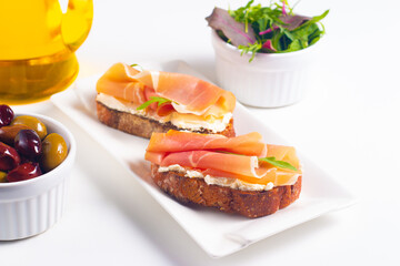 Two bruschettas with prosciutto, ricotta cheese and rucola on ciabatta bread. Olive and spices  decorated.