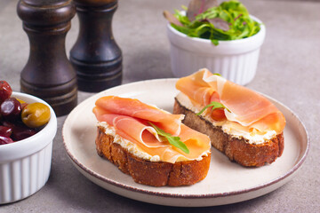 Two bruschettas with prosciutto, ricotta cheese and rucola on ciabatta bread. Olive and spices  decorated.