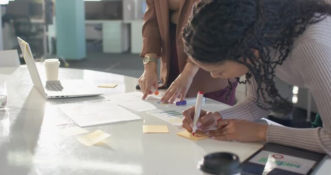 Two female colleagues in blazer at desk highlighting charts, jotting sticky notes drafting outline