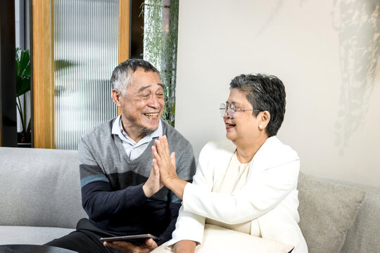 Happy retired senior Asian couple making high five hand touch while sitting on the sofa in living room at home. Smiling mature elderly lover touch each other hands with hi five to make a deal.