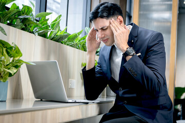 Young businessman in suit feeling bored and stressed while working on laptop computer. Upset business staff having a headache, putting head in hands during working online in green modern office.
