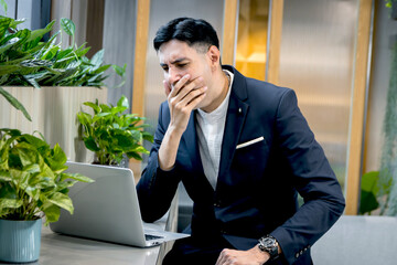 Young businessman in suit feeling bored and sleepy while working on laptop computer. Exhausted business staff yawning, want to take a nap during using a computer to work online in green modern office.