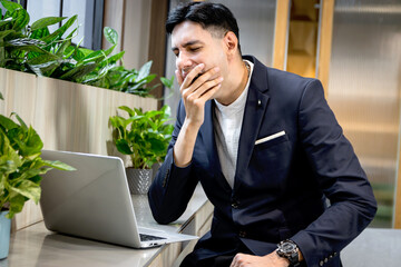 Young businessman in suit feeling bored and sleepy while working on laptop computer. Exhausted business staff yawning, want to take a nap during using a computer to work online in green modern office.