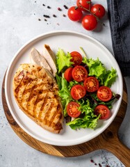 Top-down photo of grilled chicken breast with char marks, fresh greens, cherry tomatoes on white plate over wood board. Bright, clean, natural daylight, healthy meal.