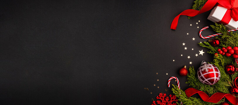 Holiday-themed flat lay featuring Christmas greenery, red and gold ornaments, gift box, candy canes, and decorative ribbon on a black background with star confetti. - Powered by Adobe