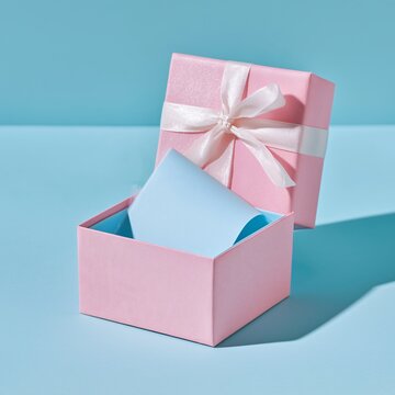 Pastel pink gift box with white ribbon is open, revealing blue card inside, set against minimalist blue background. scene conveys sense of anticipation and surprise