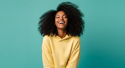 Fototapeta premium An energetic woman with voluminous afro hair and a bright smile expresses pure joy. Her yellow hoodie and the vibrant teal background create a dynamic and cheerful atmosphere.
