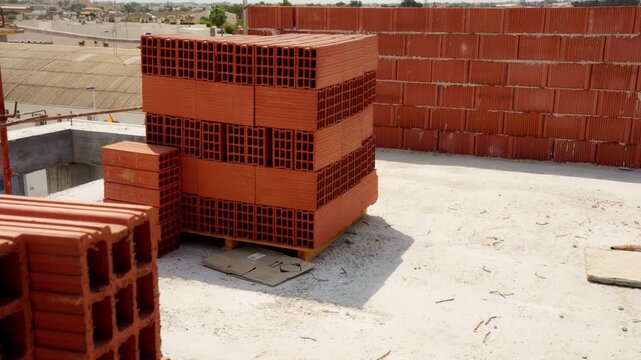 Hollow red clay bricks stacked on a wooden pallet at a sunny construction site, with a newly built wall and distant city skyline under a clear blue sky, ready for masonry work