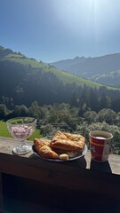 Fototapeta premium Scenic mountain breakfast with croissants, cheese, coffee, and yogurt in a glass bowl, set against a backdrop of sunlit alpine hills and forest in the early morning light.