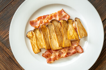 Fried eryngii mushrooms served with crispy bacon slices on white plate