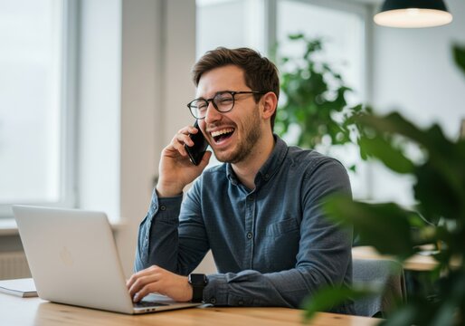 Young professional man with glasses laughing while talking on his mobile phone at his desk with a laptop - Powered by Adobe