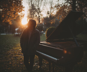 Zdjęcie pianisty stojącego przy fortepianie w plenerze, w jesiennym ogrodzie, oświetlonego zachodzącym słońcem, oddające melancholijny nastrój.