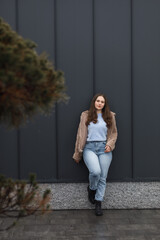 A girl in a winter parka, wearing blue jeans and a warm jacket. She has blond hair. She stands against a dark metal wall.