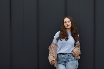 A girl in a winter parka, wearing blue jeans and a warm jacket. She has blond hair. She stands against a dark metal wall.