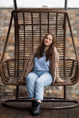 A girl in blue jeans and a warm jacket sits on a wicker bench on the terrace