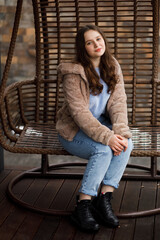 A girl in blue jeans and a warm jacket sits on a wicker bench on the terrace