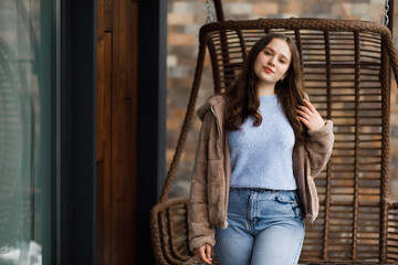 A girl in blue jeans and a warm jacket sits on a wicker bench on the terrace
