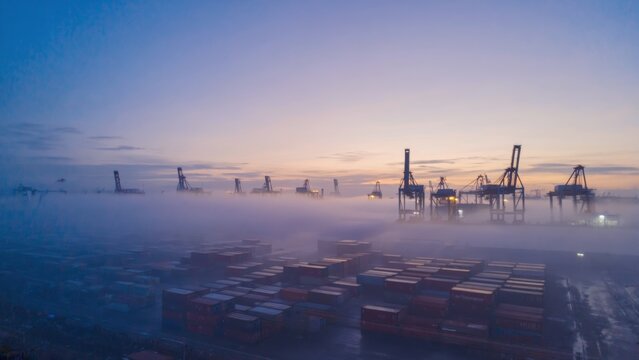 Industrial cargo port covered in morning fog with shipping containers, cranes silhouetted against twilight sky, representing global trade, maritime logistics, freight transport, import-export business