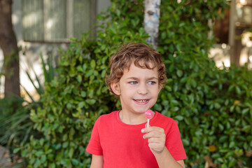 A cute and happy boy in a red t-shirt enjoys a sweet lollipop outdoors with a green leafy background