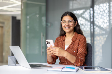 Obraz premium Young indian woman working in an office, sitting at a desk and smiling while holding a smartphone, actively engaging with technology and balancing work with communication