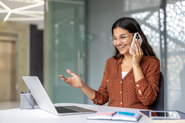 Young indian woman worker in eyeglasses having conversation on mobile phone and gesturing with hand while working on laptop in modern office environment