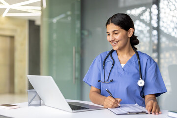 Smiling female doctor or nurse wearing a stethoscope and scrubs, sitting at a desk, typing on a laptop, and recording information on a clipboard, representing modern healthcare documentation