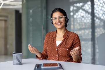 Indian woman in glasses and headset smiling, sitting at an office desk, actively engaging in an online meeting, and supporting clients via a call center