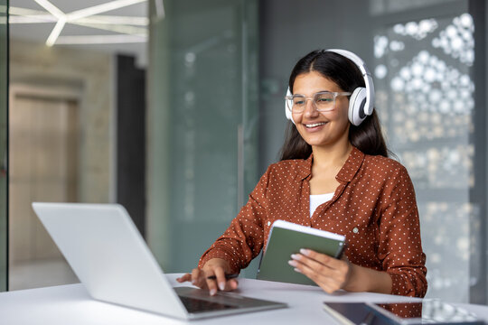 Young woman wearing headphones and glasses, happily engaging in online education or remote work, using her laptop and holding a tablet in a modern office environment - Powered by Adobe