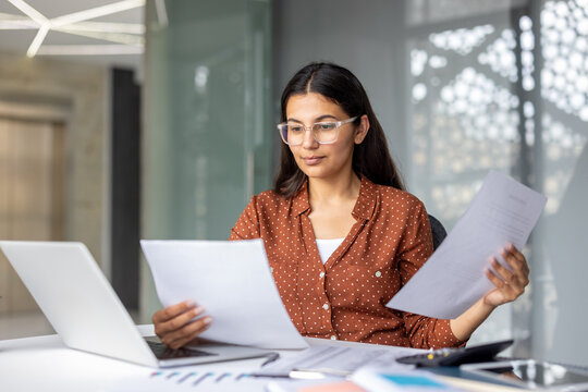 Young woman wearing glasses diligently reading and comparing financial reports and papers at her desk while working on a laptop in a contemporary office environment