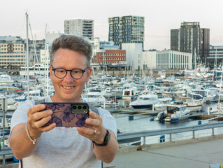 Caucasian tourist taking a selfie with his smartphone during evening in Bodø, Norway. Yachts in the harbor of Bodo in the background. Norwegian city, solo traveller taking photo with cellphone.