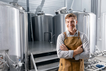 Fototapeta premium Portrait happy worker male brewer in apron on background modern brewery with fermentation tanks