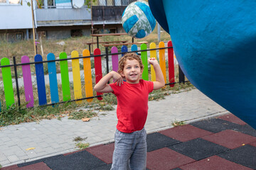 A cute little boy is holding a blue ball on a colorful playground, looking up with a thoughtful expression