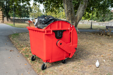 Red dumpster in park setting. This photo represents urban waste management, environment, cleanliness, and public space maintenance.