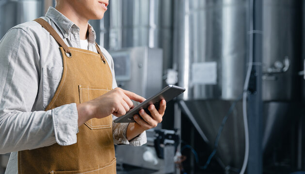 Worker brewer in apron using tablet in brewery for craft beer production