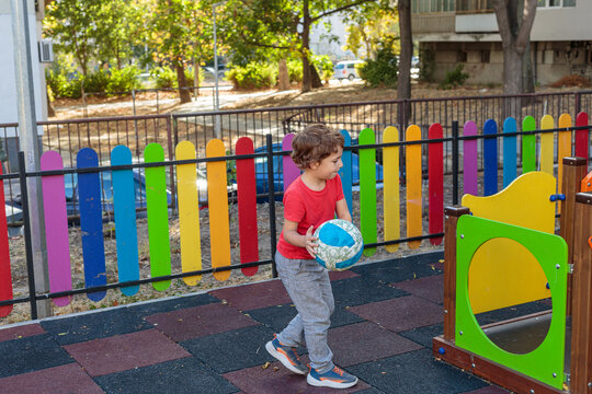A cheerful child in a red shirt running towards the camera while playing with a ball on a colorful playground