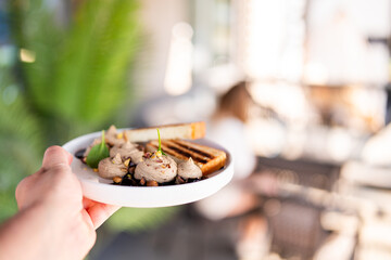 Gourmet appetizer on white plate with creamy spread, microgreens, crushed nuts, and toasted bread. Outdoor dining setting with blurred background and natural light.