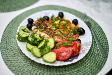Grilled fish with vibrant vegetables and olives served on white plate. Decorative green mat adds texture. Bright natural light enhances fresh ingredients colors