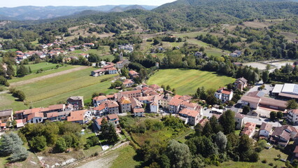 Aerial view of Battaglia, Mioglia, Italy, showcasing a charming Italian village with red-tiled roofs nestled amidst vibrant green fields and rolling forested hills under a clear September sky.