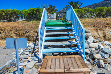 Wooden stairs from rocky beach leading to hotel on background of green bushes and high mountains. Greece. Kos.