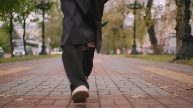 lady leg running down brick pathway in city park wearing black coat and white sneakers, autumn leaves scattered around, blurred traffic in background creating urban seasonal atmosphere