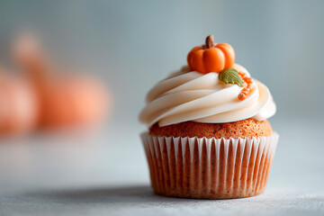 A pumpkin-topped cupcake with swirled vanilla buttercream, golden accents and a tiny leaf, set against a soft, neutral backdrop.